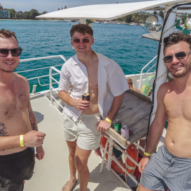 Three young men in sunglasses and swimwear stand and smile on a sunny boat deck, holding drinks. Other people and blue water are visible in the background, creating a fun, relaxed summer atmosphere.