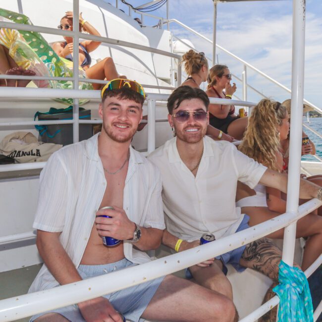 Two men in light-colored shirts sit and smile on the deck of a boat, surrounded by other people relaxing in swimwear. The sunny weather and blue sky suggest a fun day out on the water.