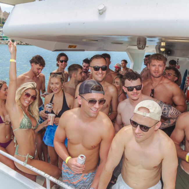 A group of young adults in swimwear smile and pose together on a boat during a sunny day, with blue water and buildings visible in the background. Some hold drinks and wear sunglasses, enjoying a lively summer party atmosphere.