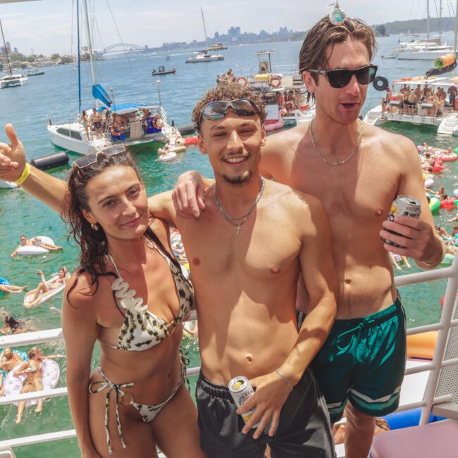 Three young adults in swimsuits pose and smile on a boat, holding drinks, with lots of people swimming and relaxing on floats in the water and several boats in the background on a sunny day.