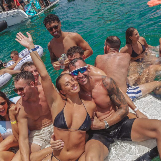 A group of young adults in swimwear laugh and pose together on a floating mat in the ocean beside yachts, enjoying a lively summer party under sunny skies.