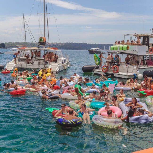 A crowd of people relax and socialize on colorful inflatable floaties in the water near several boats on a sunny day, with a scenic coastline in the background.
