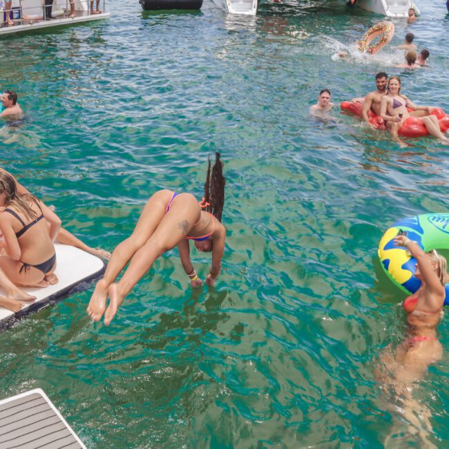 People enjoy a sunny day on the water; some relax on a floating dock, others swim or float on inflatables. One woman dives into the water, while yachts are anchored in the background.