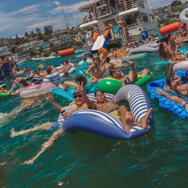 A large group of people float on colorful inflatables and swim in a crowded, lively pool party near boats, enjoying sunny weather and festive atmosphere.