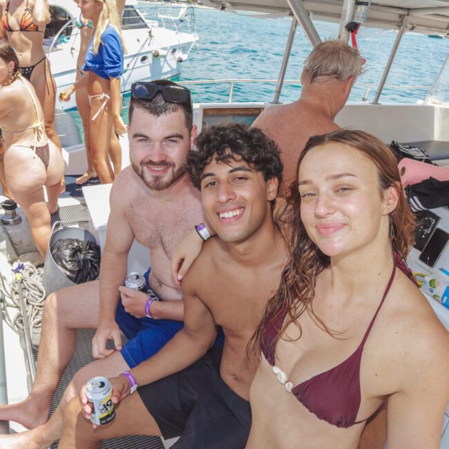 Three young adults in swimsuits smile at the camera while sitting on a boat with others in the background. The sunny scene shows clear blue water and another boat nearby, creating a lively, summery atmosphere.