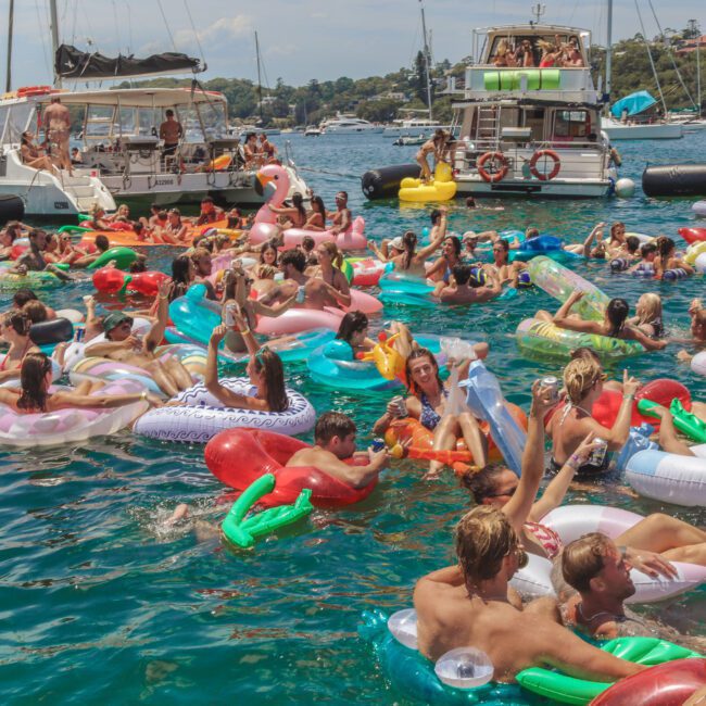A large group of people on colorful inflatable pool floats gather in the water near several boats on a sunny day, enjoying a lively summer party.