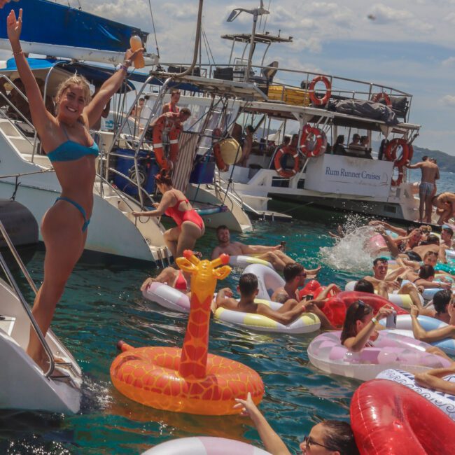 People relax on colorful inflatable floats in the water near boats on a sunny day. A smiling woman in a blue bikini poses on a yacht, while others lounge and swim around her, creating a lively, festive scene.