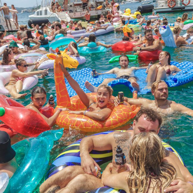 A crowded pool party with many people lounging on colorful inflatable floats, swimming, and relaxing in the water. One person in the center excitedly poses and makes a peace sign among the lively scene.