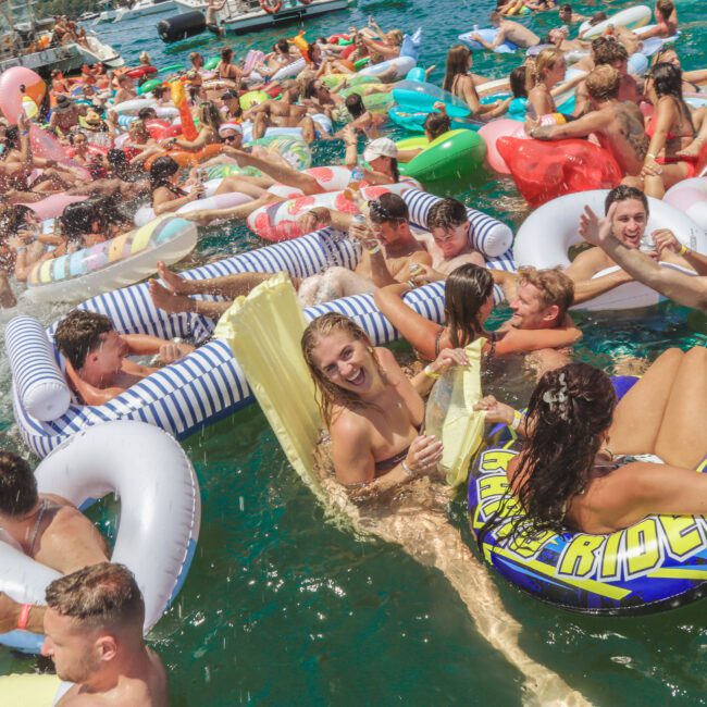 A large group of people in swimsuits enjoy a lively pool party, floating on colorful inflatables in bright blue water under sunny skies. Everyone looks cheerful and relaxed, laughing and celebrating together.