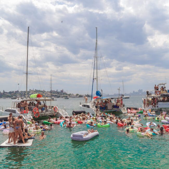 A large group of people on colorful inflatables and in the water gather between boats in a sunny harbor. Catamarans and a party boat are anchored nearby under a partly cloudy sky, with a city skyline in the background.