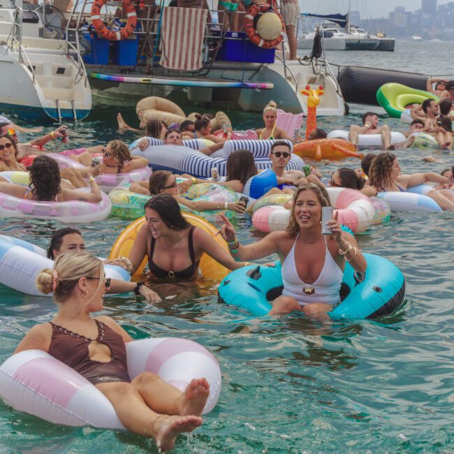 A large group of people in colorful swimsuits relax and laugh on inflatable pool floats in the water near a yacht, with a city skyline visible in the background. The atmosphere is lively and fun.