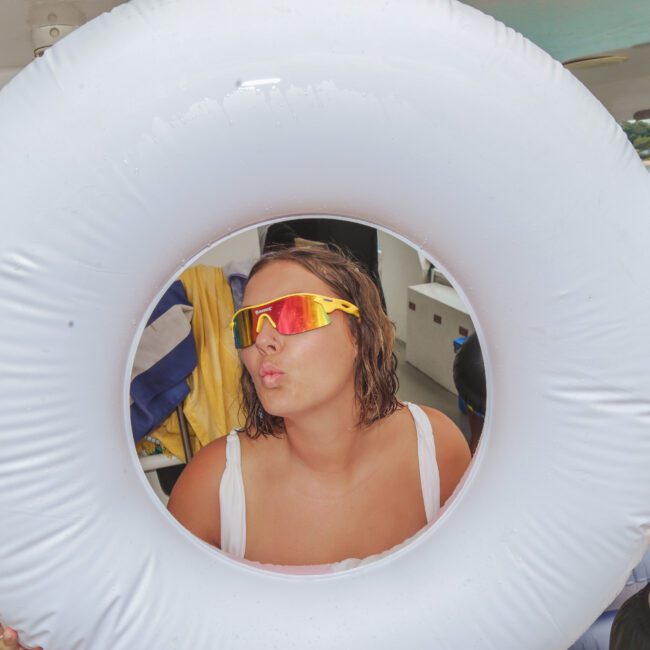 A woman wearing reflective sunglasses poses playfully, puckering her lips through the center of a white inflatable ring on a boat, with the ocean and shoreline visible in the background.