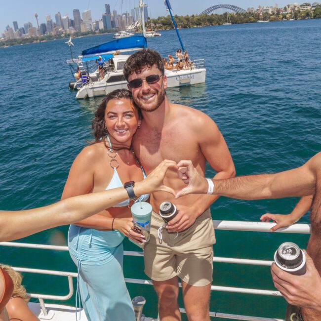 A group of smiling young adults in swimwear enjoy drinks on a boat, with a city skyline, harbor, and bridge in the background on a sunny day.