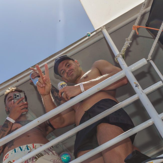 Two men stand shirtless on a boat deck behind railings; one holds a phone and the other flashes a peace sign while holding a drink. Bright sunlight highlights the scene, with part of the boat structure visible.