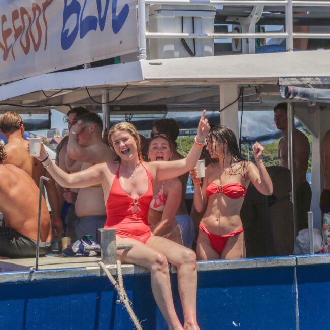A group of young adults in swimsuits are enjoying a party on a boat in sunny weather. Two women in the foreground smile and gesture energetically, while others socialize in the background. A sign reads "Barefoot Blue.