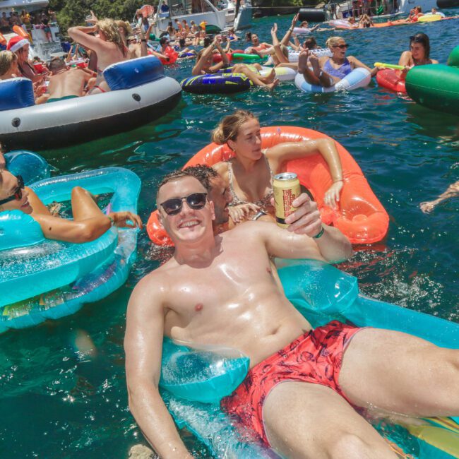 A man in red swim trunks smiles and holds a drink while lounging on a blue pool float in a crowded, lively pool party with people on various colorful inflatables in the water around him.