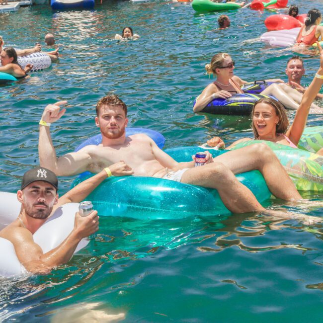 A group of people float on colorful inflatable tubes in a sunny, crowded lake. Three friends in the foreground smile and gesture at the camera, holding drinks. Other groups relax and mingle in the background.