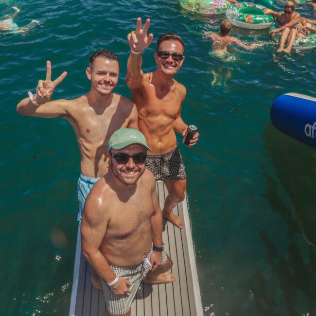 Three men in swimwear pose and smile on a dock by the water, flashing peace signs. Other people relax on colorful pool floats in the background. It’s a sunny day with a lively, fun atmosphere.