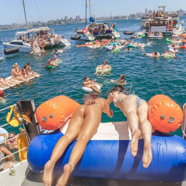 Two women in swimsuits lie on a float at the edge of a boat, facing a crowded scene of people on boats and inflatables enjoying a sunny day on the water, with a city skyline in the background.