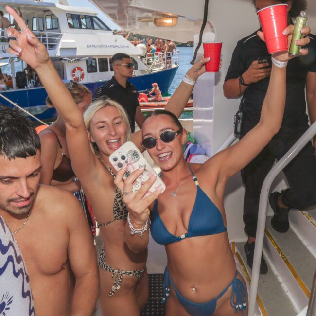 A group of young people in swimsuits celebrate on a boat, smiling, holding drinks, and taking a selfie. Other boats and people are visible in the background, with a festive summer atmosphere.