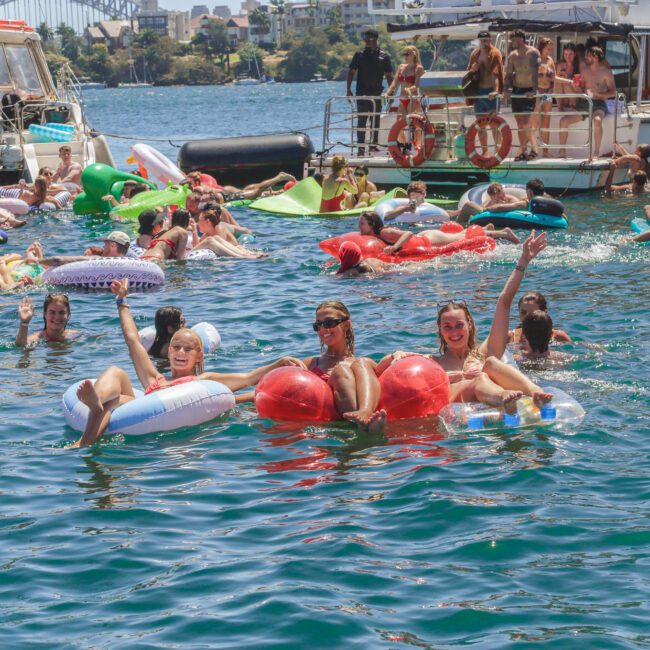 A group of people relax and have fun on colorful inflatable floaties in a lively harbor, surrounded by boats and others enjoying the sunny day on the water.