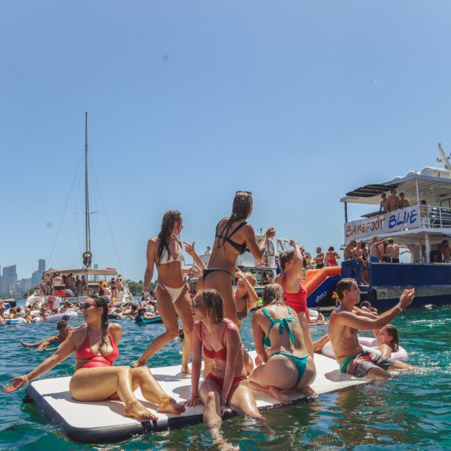A group of people in swimsuits sit and stand on a floating mat in the ocean, surrounded by boats and others in the water, with a clear blue sky and city skyline in the background.