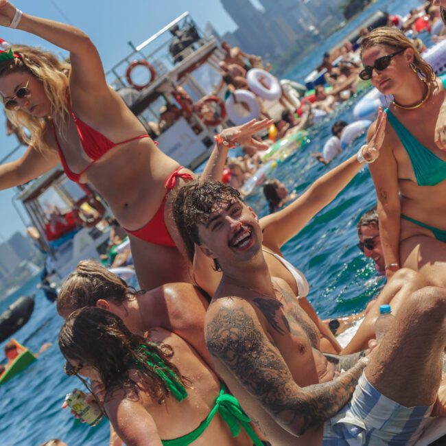 A group of young adults in swimsuits enjoy a lively boat party on the water, dancing, laughing, and socializing under the sun with city skyscrapers visible in the background.