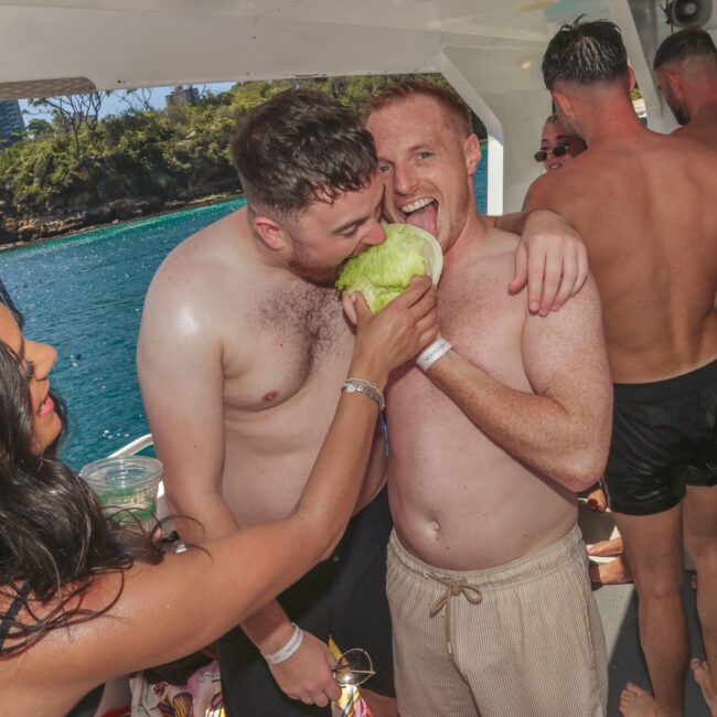 Two shirtless men on a boat laugh as they bite into the same fruit, held by a woman. Other people in swimwear are in the background, with the ocean and trees visible outside. A “Mind your head” sign is overhead.