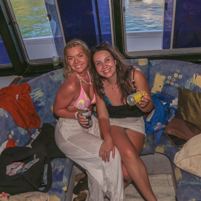 Two women smiling and sitting on a patterned couch on a boat, each holding a drink. The area is cozy with cushions, bags, and a window showing water outside. The atmosphere looks relaxed and cheerful.