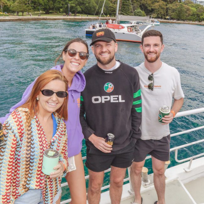 Four smiling young adults pose together on a boat holding drinks, with clear blue water, another boat, and lush green trees visible in the background under a bright sky.