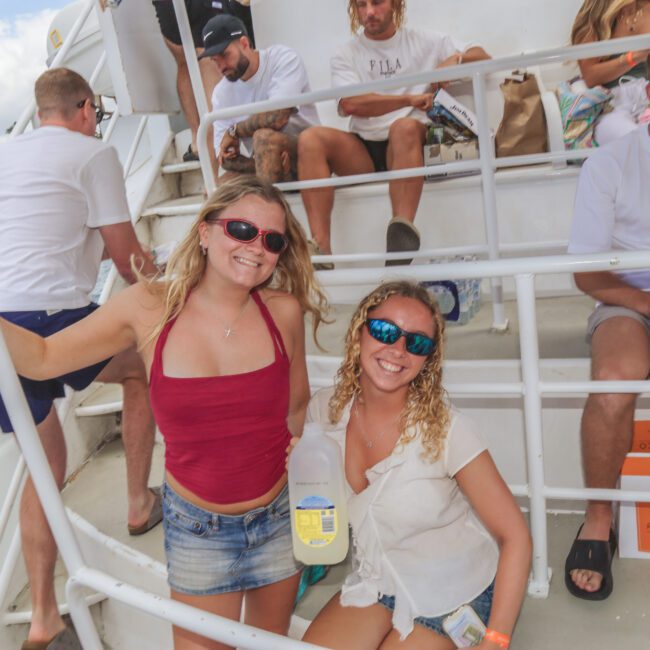 Two smiling women in sunglasses pose on a boat. One holds a large bottle of lemonade. Other people sit around them, some with drinks. The background shows water and trees under a partly cloudy sky.