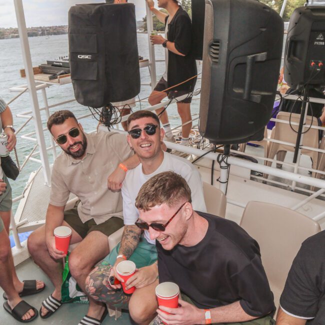 A group of smiling men in casual summer clothes sit together holding cups on a boat, enjoying a sunny day on the water with speakers and more people in the background.