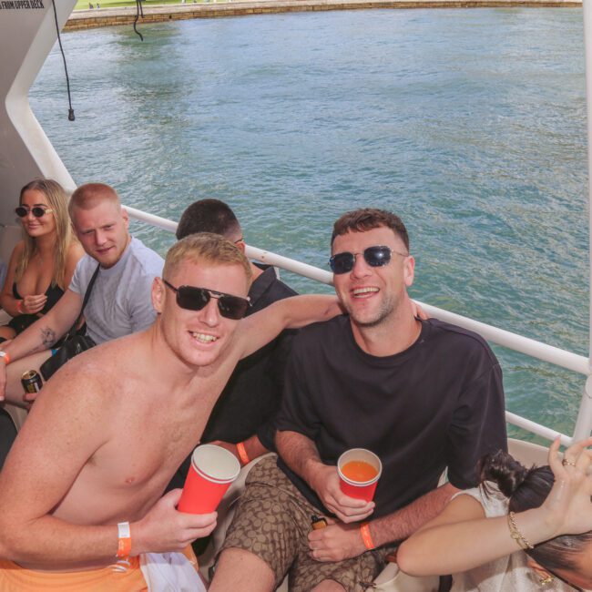 A group of people sit and smile on a boat under sunny weather. Two men in front hold red cups and pose for the camera, others relax nearby. Water and another boat are visible in the background.