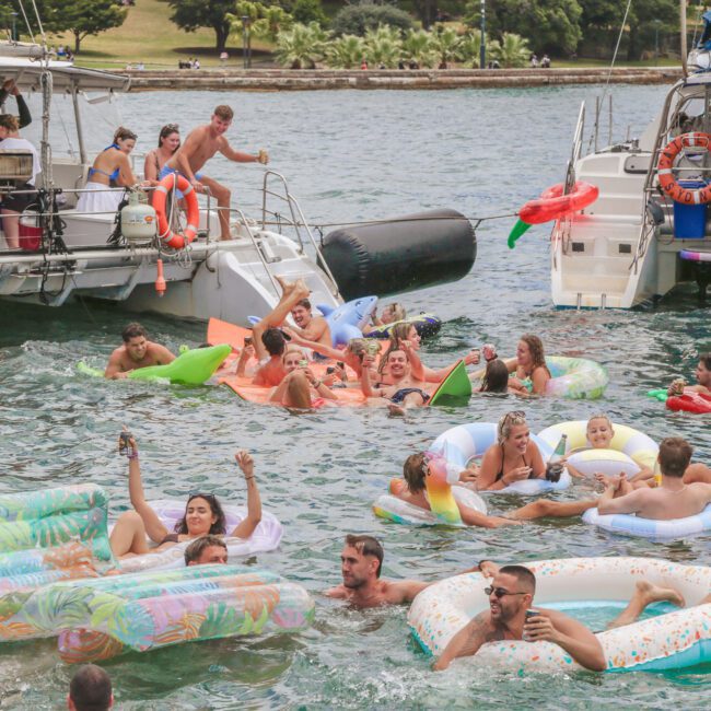 A group of people relax and have fun on colorful pool floats in the water between two docked boats, enjoying a sunny day near a grassy shoreline with palm trees.