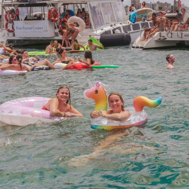 People are swimming and relaxing on colorful inflatables in the water near yachts. Two women in the foreground are smiling, one on a unicorn float and the other on a donut float, with the Sydney Opera House in the background.
