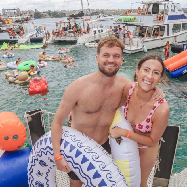 A smiling man and woman in swimsuits hold inflatable pool floats on a dock, surrounded by people swimming and relaxing on inflatables, with boats and a city skyline in the background.