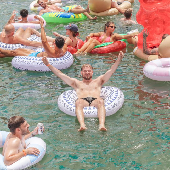 A group of people relax on colorful inflatable tubes and floats in a sunny body of water. A smiling man in the foreground raises his arms while others socialize and float nearby.