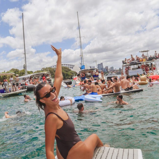 A smiling woman in a dark swimsuit sits on a boat's swim platform, waving at the camera. Behind her, many people are swimming and relaxing on floats in the water at a lively boat party under a partly cloudy sky.