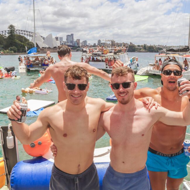 Three men in swim trunks, sunglasses, and holding drinks smile with arms around each other at a lively boat party on the water. Other people, boats, and city skyline are visible in the background under partly cloudy skies.
