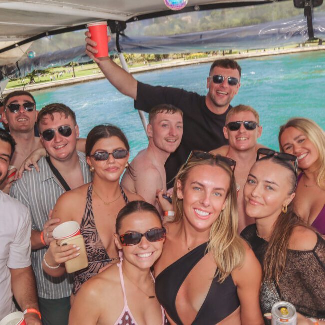 A group of people in swimsuits and sunglasses smiling and holding drinks on a boat, with blue water and green trees visible in the background. It appears to be a sunny day and everyone looks happy.