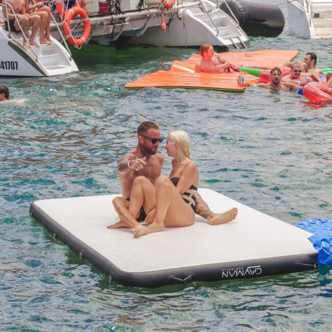 A man and woman sit together on a floating white platform in the water, surrounded by people swimming and relaxing on inflatables near docked boats on a sunny day.