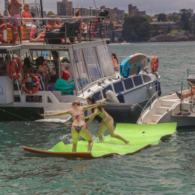 Two women in bikinis balance playfully on a floating mat between two boats, with people watching and relaxing on the boats. The scene is lively and set on a sunny day near the shore, city buildings visible in the background.