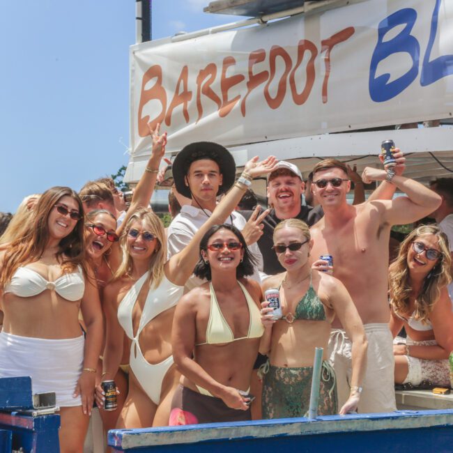 A group of people in swimsuits smile and pose with drinks at an outdoor pool party under a "Barefoot Blue" banner on a sunny day.