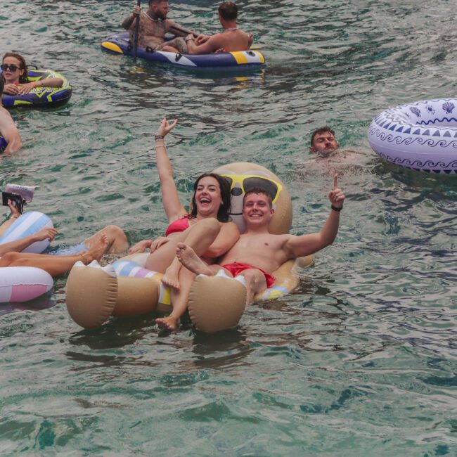A group of young adults float on inflatable pool toys, smiling and giving thumbs up in a large body of water, surrounded by others on various floaties. The atmosphere is lively and carefree.