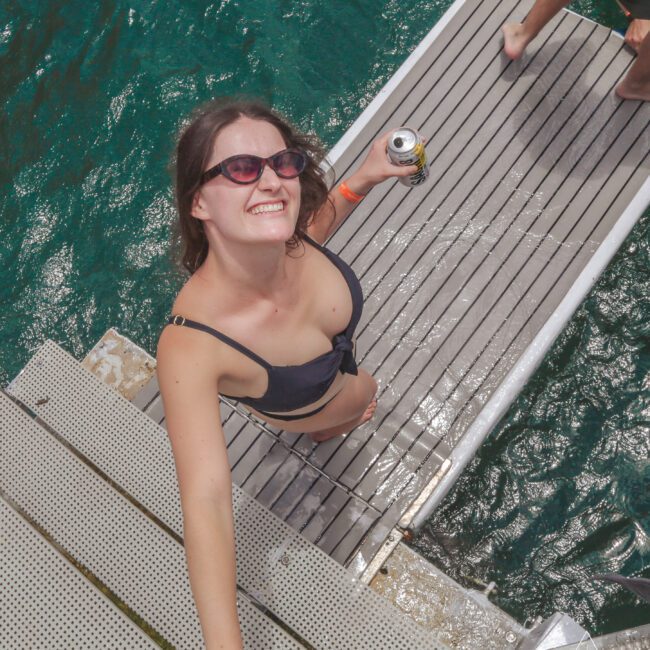 A smiling woman in a black bikini and sunglasses holds a can of drink while standing on a dock over blue-green water. She looks up toward the camera, enjoying a sunny day. Another person's leg is partially visible in the corner.
