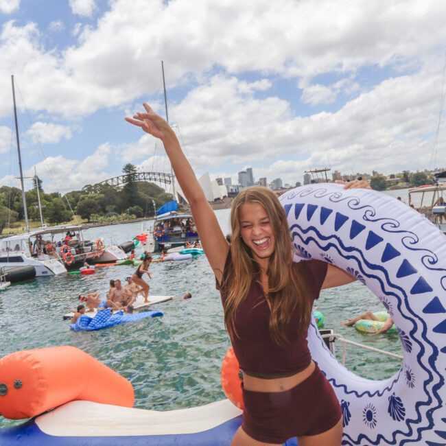 A smiling woman in a maroon outfit holds a large inflatable ring on a crowded waterfront, surrounded by boats and people swimming and relaxing on floats under a cloudy sky.
