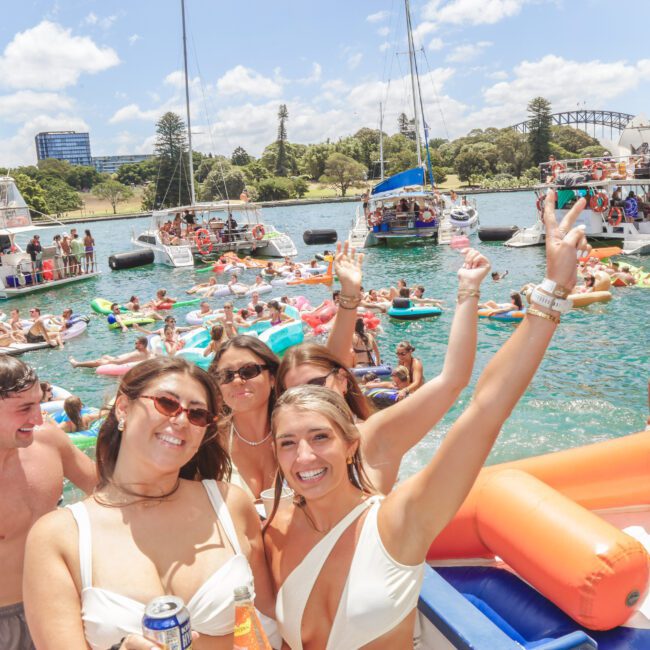 People in swimsuits enjoying a lively boat party on a sunny day, with inflatable rafts and yachts on the water, city skyline and Sydney Harbour Bridge visible in the background. Smiling guests pose and raise their arms.
