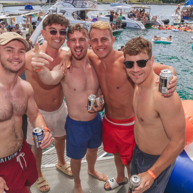 Five smiling young men in swimwear stand on a boat holding drinks, with yachts and people floating on inflatables in the water behind them under sunny skies.