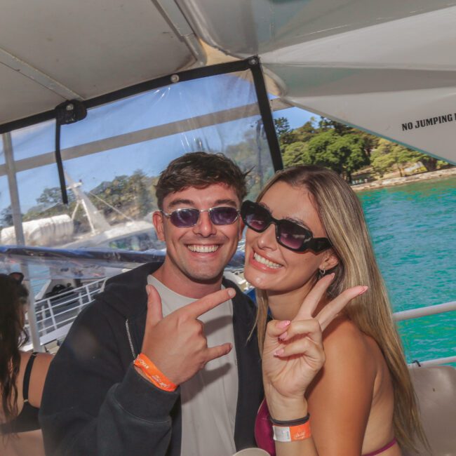 A smiling man and woman wearing sunglasses pose for the camera on a boat. The woman flashes a peace sign, and both wear orange wristbands. Turquoise water and trees are visible in the background.
