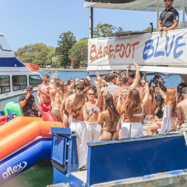 A group of people in swimwear are partying on a boat called "Barefoot Blue," dancing and socializing under sunny skies. Inflatable pool floats are in the water nearby, and another boat is visible in the background.